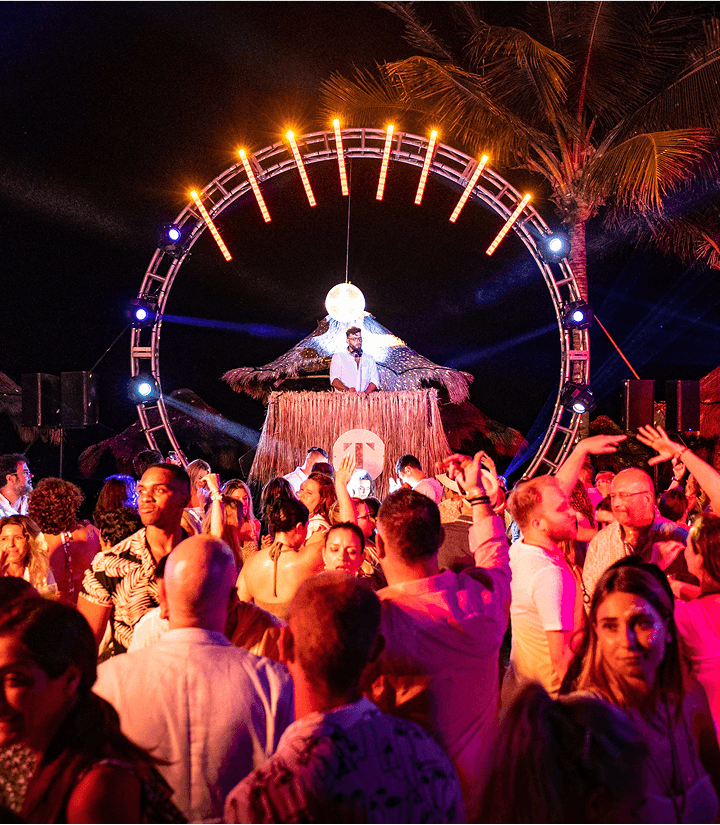 A crowd dancing on the beach in front of a DJ booth under a ring of lights